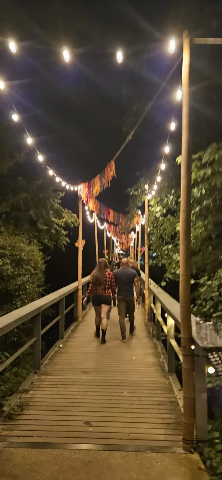 Twee bezoekers lopen in de avond over een houten loopbrug met lampjes en oranje slingers