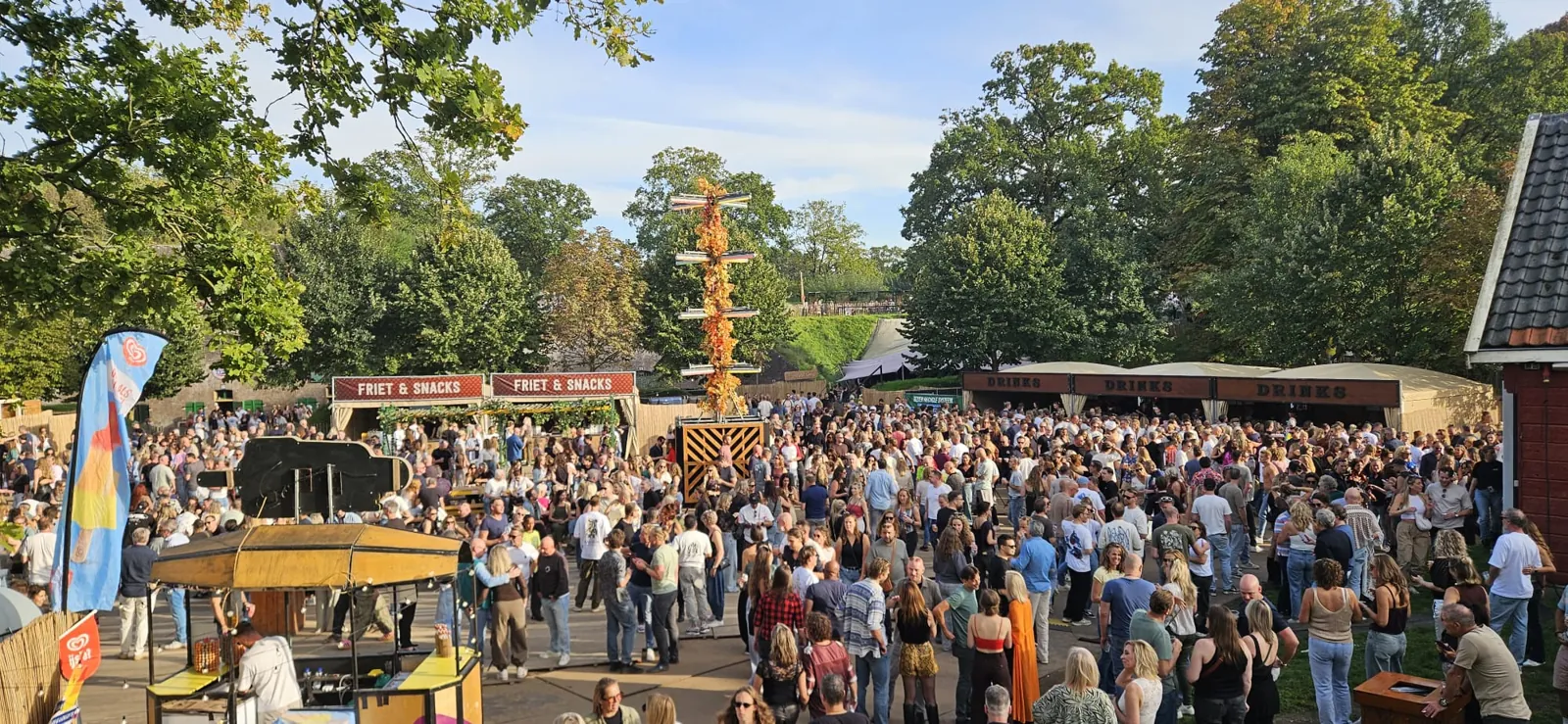 Festivalplein overdag met kraampjes voor friet en drankjes, een versierde mast met oranje bladeren en veel bezoekers