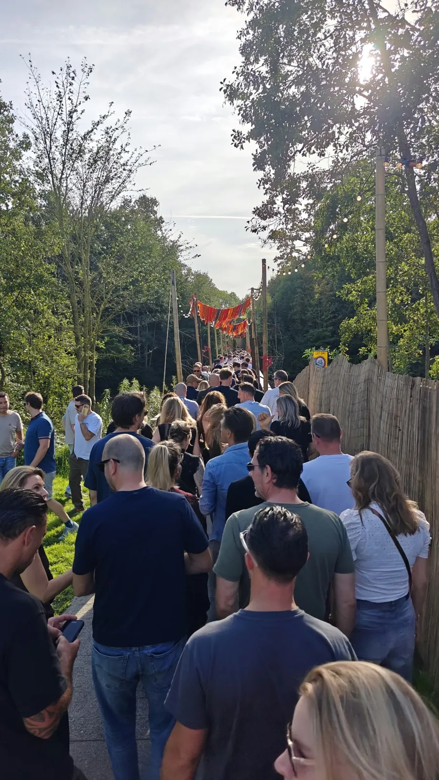 Lange stroom bezoekers op een pad onder oranje slingers tussen de bomen, op weg naar het festivalterrein