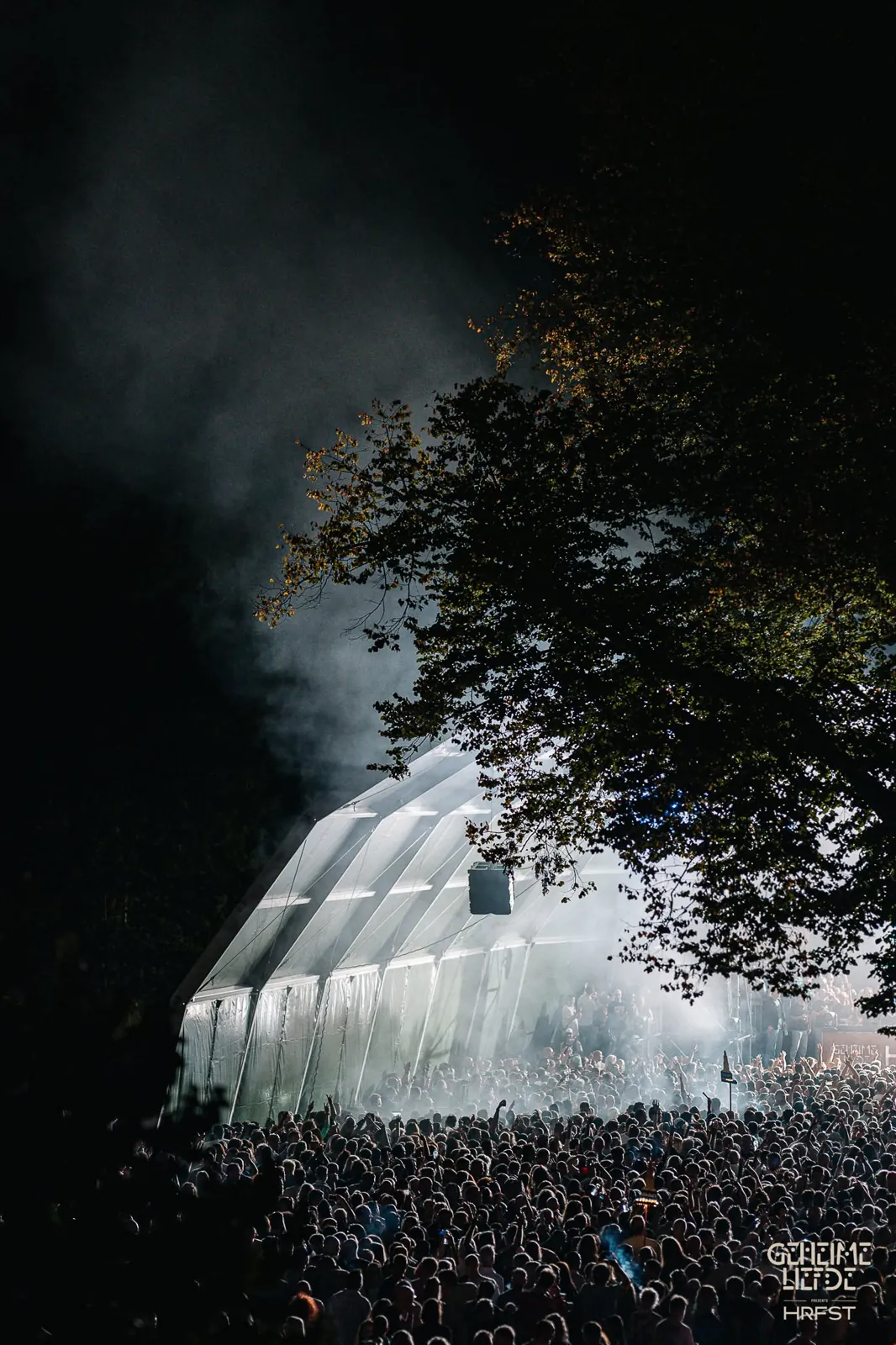 Verlichte tent in de nacht, deels verborgen achter een boom, met rook en een volle menigte ervoor