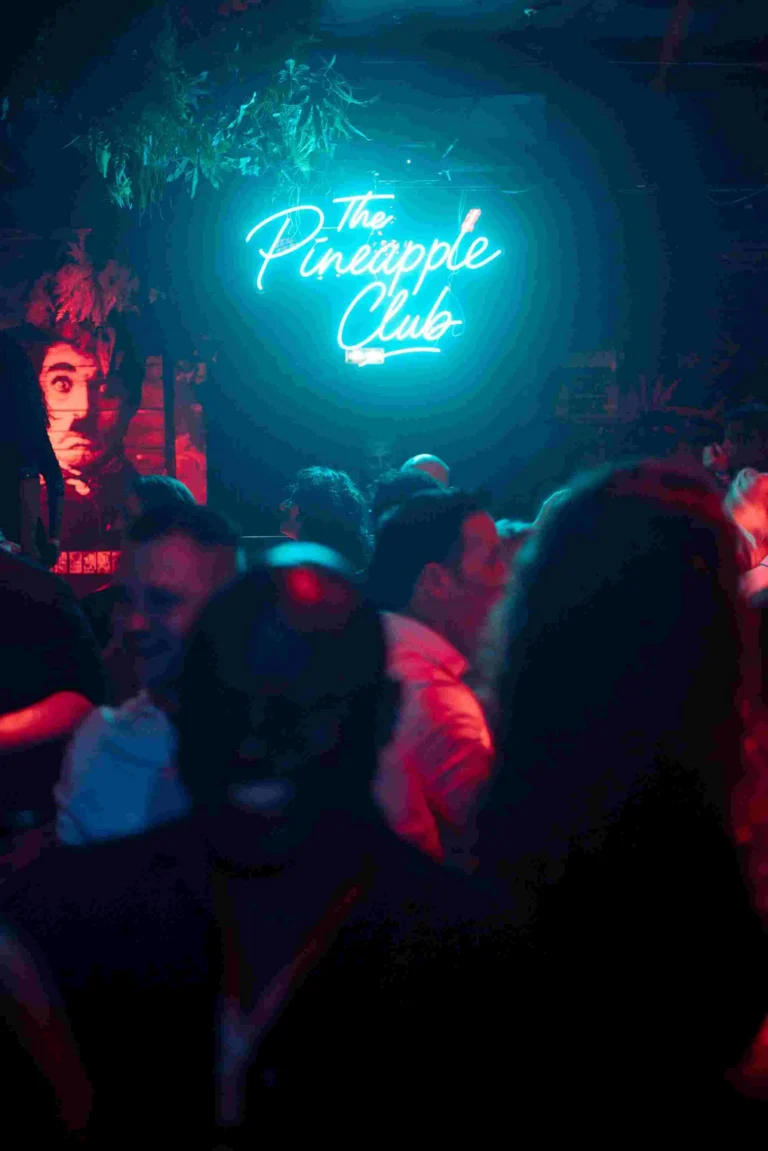 Het neonbord ‘The Pineapple Club’ gloeit boven een levendige, ingetogen verlichte dansvloer.