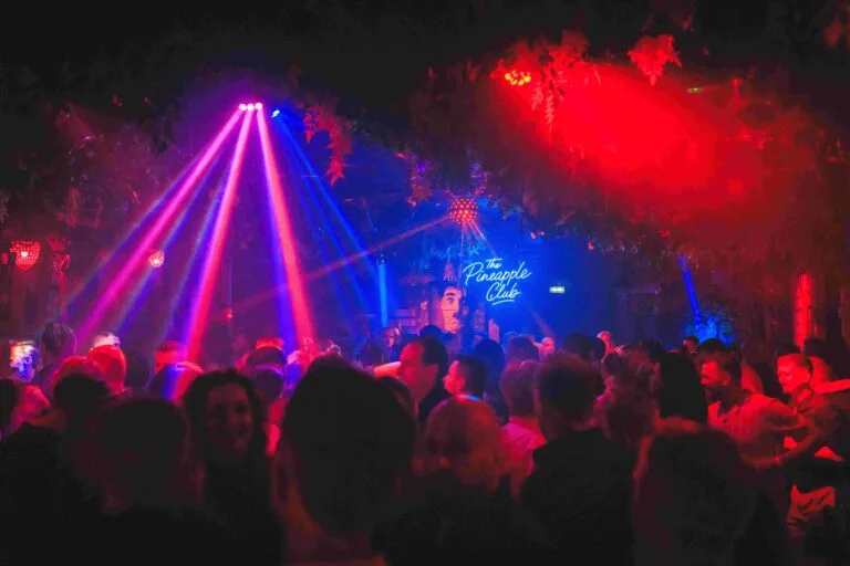 Kleurrijke lichtstralen boven een volle dansvloer, met het Pineapple Club-bord op de achtergrond.